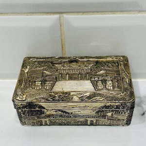 May include: A rectangular silver-colored box with detailed relief carvings. The lid features a prominent architectural scene, while the sides display dragon motifs and landscape elements. The box is likely made of metal and has a vintage appearance.