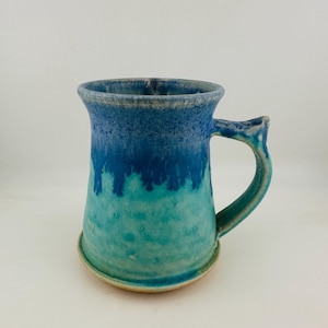 May include: A ceramic mug featuring a two-tone glaze with a deep blue upper section that transitions into a lighter turquoise base. The mug has a wide, flared rim and a sturdy, curved handle attached to the side, with a visible light tan clay base.
