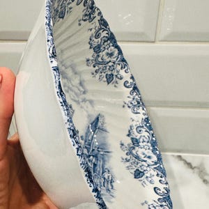 May include: A white ceramic bowl with a scalloped edge and a blue transferware pattern featuring floral motifs and a detailed landscape scene with figures. The interior surface is decorated with intricate blue illustrations on a white background.