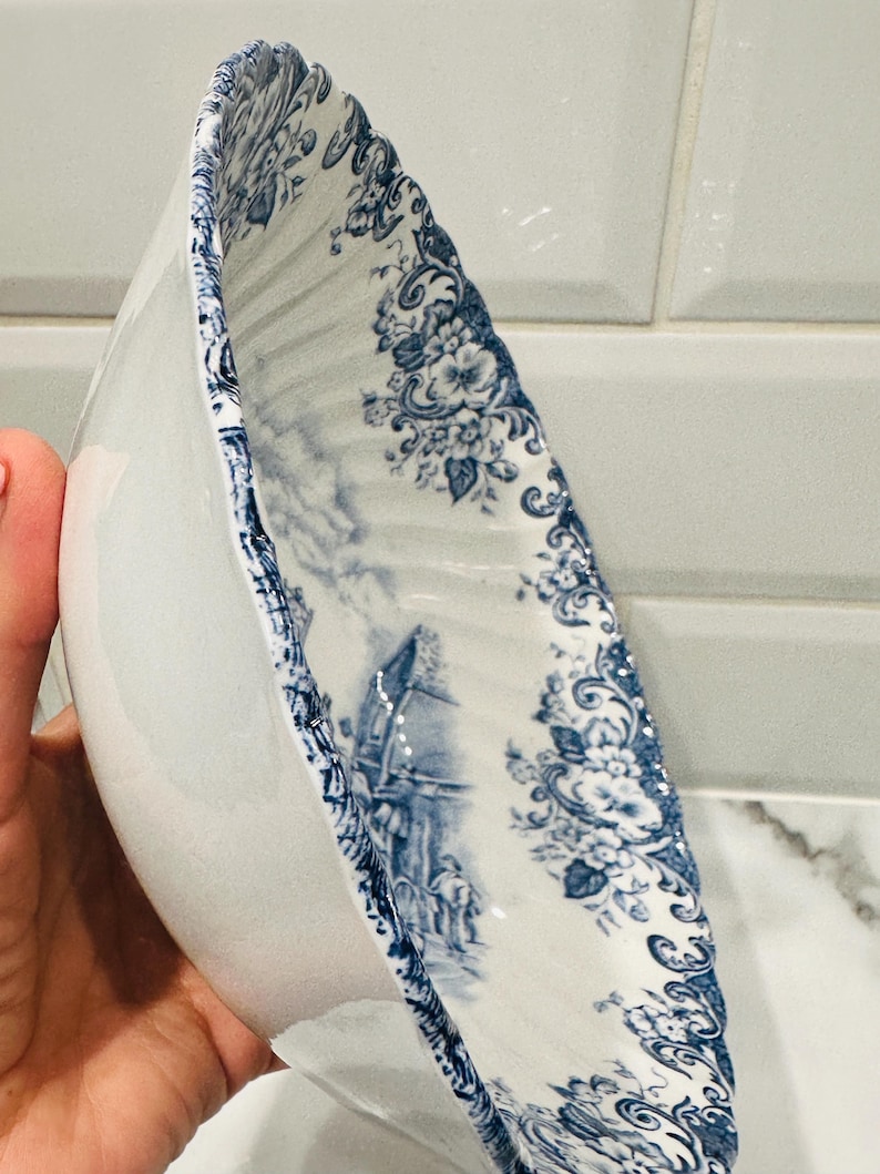 May include: A white ceramic bowl with a scalloped edge and a blue transferware pattern featuring floral motifs and a detailed landscape scene with figures. The interior surface is decorated with intricate blue illustrations on a white background.