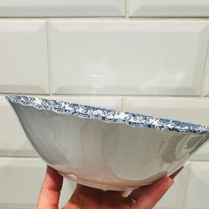 May include: A ceramic bowl with a smooth white interior and a scalloped rim featuring a detailed blue and white floral pattern. The exterior of the bowl is plain white, and the overall shape is wide and shallow with a curved, elegant profile.