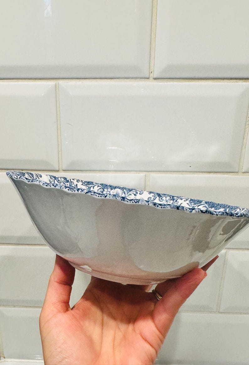 May include: A ceramic bowl with a smooth white interior and a scalloped rim featuring a detailed blue and white floral pattern. The exterior of the bowl is plain white, and the overall shape is wide and shallow with a curved, elegant profile.