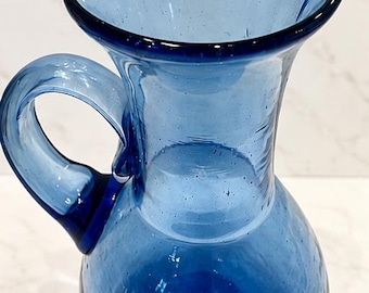 May include: A hand-blown cobalt blue glass pitcher featuring a rounded base, a slender neck, and a wide, flared rim with a small pouring spout. The glass has a translucent quality with visible air bubbles throughout the structure.