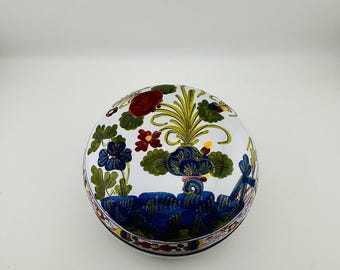 Könnte beinhalten: Runde, weiße Keramik-Schmuckdose mit Deckel. Die Dose ist mit einem Blumendesign mit blauen, roten und grünen Blumen und Blättern verziert. Der Rand der Dose hat einen dekorativen Rand.