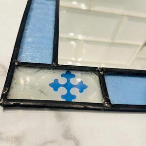 K&ouml;nnte beinhalten: Ein rechteckiger Spiegel, der mit Buntglas-Paneelen in Hellblau und Wei&szlig; gerahmt ist. Der Rahmen hat eine schwarze Metalll&ouml;tung und enth&auml;lt dekorative wei&szlig;e Glasfliesen mit einem blauen, kreuzf&ouml;rmigen Emblem-Muster.
