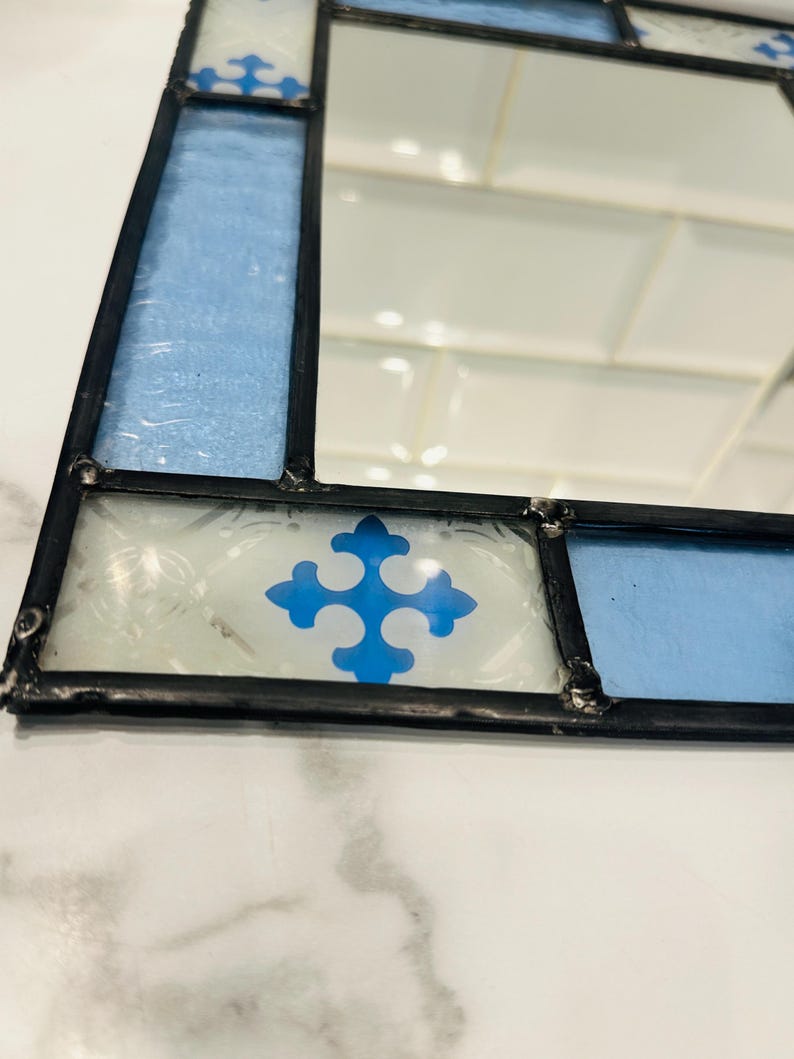 K&ouml;nnte beinhalten: Ein rechteckiger Spiegel, der mit Buntglas-Paneelen in Hellblau und Wei&szlig; gerahmt ist. Der Rahmen hat eine schwarze Metalll&ouml;tung und enth&auml;lt dekorative wei&szlig;e Glasfliesen mit einem blauen, kreuzf&ouml;rmigen Emblem-Muster.