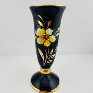 Op de afbeelding: Zwarte vaas met gouden rand en voet. Een geel en witte bloem met een rood hart is op de voorkant geschilderd. Goudkleurige accenten sieren de bloem en de stengel. De vaas is ongeveer 18 cm hoog.