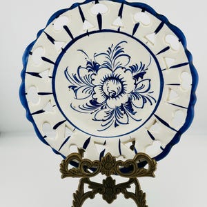Puede incluir: Un plato decorativo blanco con un diseño floral azul y un borde azul festoneado. El plato tiene formas recortadas alrededor del borde y se exhibe en un soporte de metal color bronce. Plato decorativo de estilo vintage.