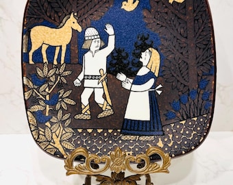Könnte beinhalten: Quadratischer Keramikteller mit einer Volkskunstszene, die einen Mann, eine Frau, ein Pferd und Vögel in einer Waldlandschaft zeigt. Der Teller ist braun mit blauen und goldenen Akzenten, präsentiert auf einem dekorativen Messingständer.