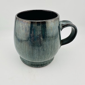 May include: A ceramic mug with a rounded body and a sturdy handle. The exterior features a reactive glaze with blended shades of deep blue, teal, and charcoal gray, creating a variegated, streaked appearance. The interior is a solid, glossy black.