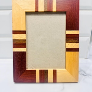 May include: A rectangular wooden picture frame with a natural wood finish and alternating stripes of light and dark wood. The frame has a simple, clean design, with a neutral-colored backing.