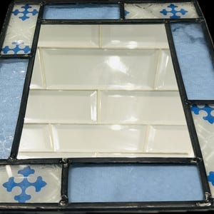 K&ouml;nnte beinhalten: Ein rechteckiger Spiegel mit einem Buntglasrahmen, der aus abwechselnden Paneelen aus hellblauem, strukturiertem Glas und wei&szlig;em Glas mit blauen kreuzf&ouml;rmigen Mustern besteht, die durch dunkle Metalll&ouml;tlinien zusammengehalten werden.