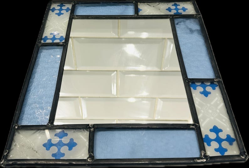 K&ouml;nnte beinhalten: Ein rechteckiger Spiegel mit einem Buntglasrahmen, der aus abwechselnden Paneelen aus hellblauem, strukturiertem Glas und wei&szlig;em Glas mit blauen kreuzf&ouml;rmigen Mustern besteht, die durch dunkle Metalll&ouml;tlinien zusammengehalten werden.