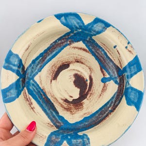 Pode incluir: Uma tigela de cerâmica redonda com uma base de cor creme, apresentando um padrão de cruz azul pintado à mão e um centro salpicado de castanho escuro. A tigela tem uma borda larga e um acabamento vidrado liso com pinceladas visíveis.