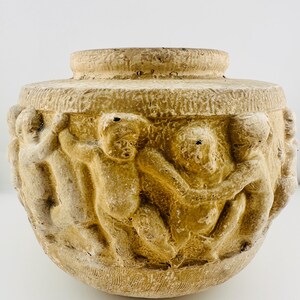 May include: A round, beige-colored vessel with a textured surface featuring a continuous relief carving of several figures holding hands in a circle around the exterior. The pot has a wide, flat rim and a rounded base with a weathered finish.