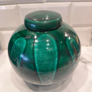 Op de afbeelding: Een ronde, groene keramische pot met een bijpassende deksel. De pot heeft een glanzende afwerking en een geribbeld ontwerp. Het deksel zit bovenop de pot en creëert een samenhangend en decoratief stuk. De pot heeft een levendige groene tint.