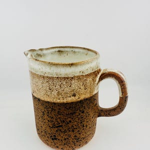 May include: A ceramic pitcher featuring a two-tone glaze with a speckled dark brown base and a smooth, creamy white upper section. The vessel includes a small pouring spout and a sturdy, curved handle that matches the speckled brown finish.