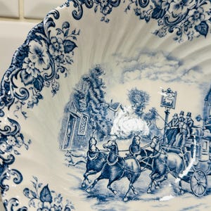 May include: A white ceramic bowl with a scalloped edge featuring a blue transferware pattern. The design depicts a horse-drawn stagecoach traveling past a building, with floral motifs decorating the rim and a detailed landscape scene in the center.
