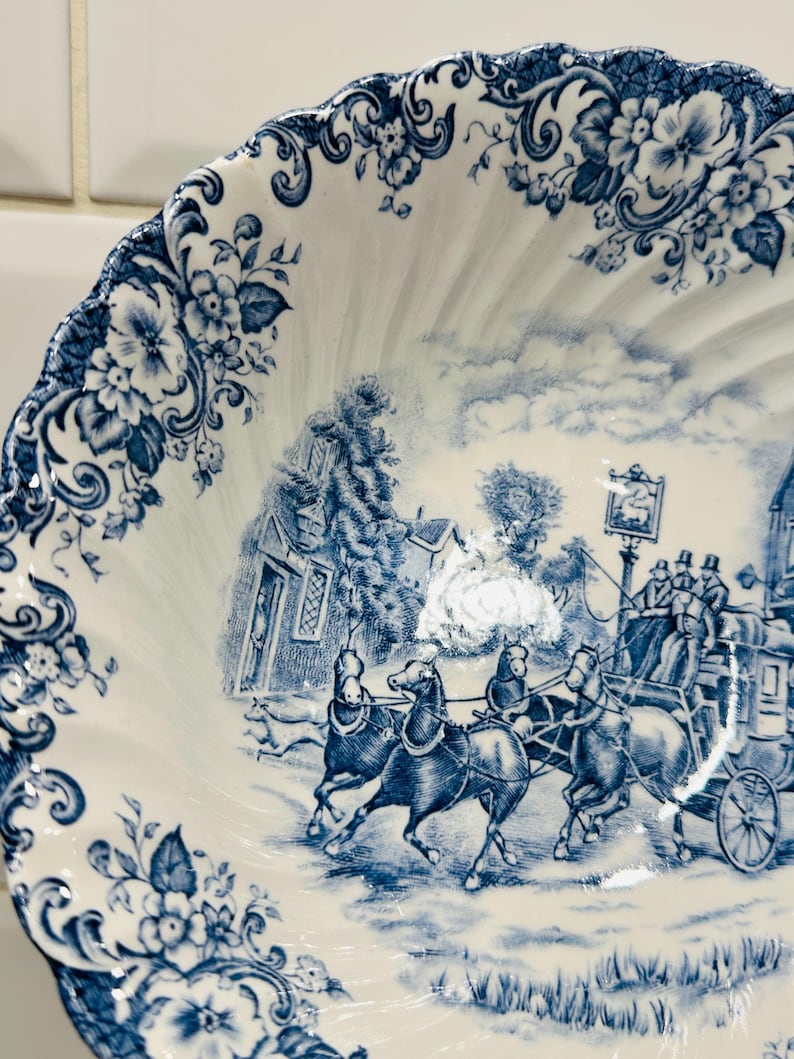 May include: A white ceramic bowl with a scalloped edge featuring a blue transferware pattern. The design depicts a horse-drawn stagecoach traveling past a building, with floral motifs decorating the rim and a detailed landscape scene in the center.