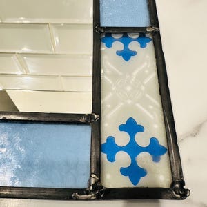 K&ouml;nnte beinhalten: Ein rechteckiger Spiegel mit einem dekorativen Rahmen aus Buntglas. Der Rahmen besteht aus hellblauen Glassegmenten und wei&szlig;en Glaspaneelen mit einem blauen kreuzf&ouml;rmigen Muster, die durch dunkle Metalll&ouml;tstellen verbunden sind.