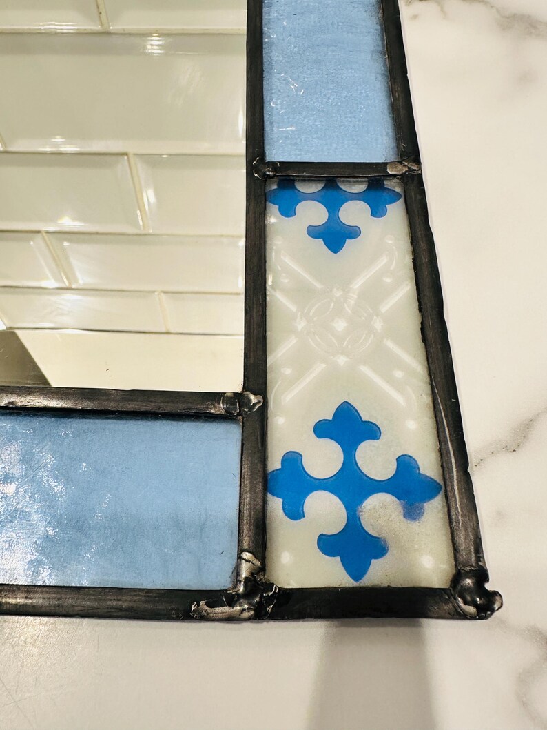 K&ouml;nnte beinhalten: Ein rechteckiger Spiegel mit einem dekorativen Rahmen aus Buntglas. Der Rahmen besteht aus hellblauen Glassegmenten und wei&szlig;en Glaspaneelen mit einem blauen kreuzf&ouml;rmigen Muster, die durch dunkle Metalll&ouml;tstellen verbunden sind.