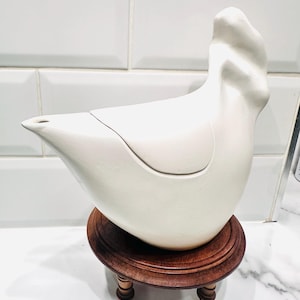 May include: A white ceramic bird-shaped ornament with a lid, displayed on a small, round, dark brown wooden stand. The stand features four short, turned legs. The bird has a smooth, matte finish.