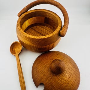 May include: A round wooden bucket with a curved handle, a matching circular lid featuring a small knob and a cutout for a utensil, and a small wooden spoon. The set displays a natural light brown wood grain pattern throughout the entire surface.