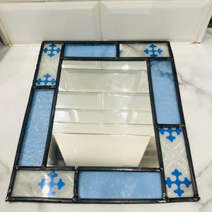 K&ouml;nnte beinhalten: Ein rechteckiger Spiegel mit einem Rahmen aus Buntglas. Der Rahmen besteht aus abwechselnd hellblau strukturierten Glasst&uuml;cken und klaren Glaselementen mit einem blauen Kreuzmuster. Die Glasteile sind durch dunkle Metalln&auml;hte verbunden.
