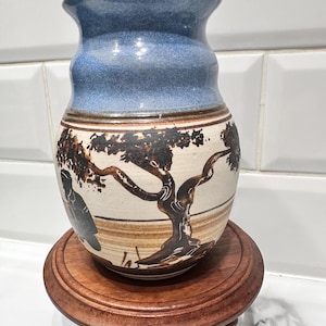 May include: A ceramic vase with a blue top and a cream-colored body featuring a brown tree and figure design. The vase sits on a small, round, wooden stand. The vase has a flared rim and a slightly textured surface.