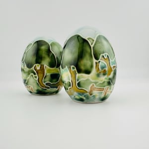 May include: Two egg-shaped ceramic sculptures featuring a glossy finish with a hand-painted landscape design. The surface displays shades of forest green, sage, and earthy brown, separated by raised white outlines that define tree and terrain shapes.