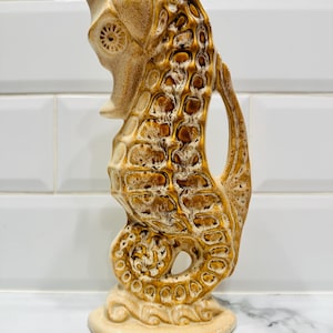 Könnte beinhalten: Keramik-Seepferdchen-Figur in Beige- und Brauntönen. Das Seepferdchen hat einen strukturierten, gemusterten Körper und einen gebogenen Schwanz. Die Figur steht auf einem runden Sockel und hat einen griffartigen Ansatz.