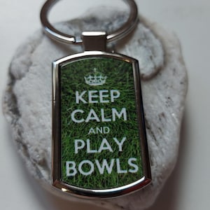 Könnte beinhalten: Ein silberner Schlüsselanhänger mit grünem Hintergrund und dem Text "Keep Calm and Play Bowls" in Weiß.