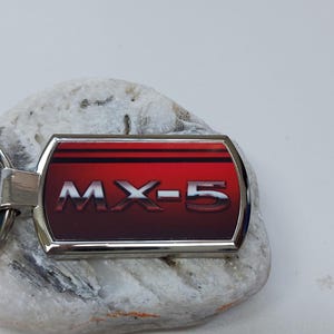 Op de afbeelding: Een zilveren sleutelhanger met een rechthoekig rood en zilveren label met daarop "MX-5" in witte letters.