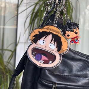 Könnte beinhalten: Ein Schlüsselanhänger mit zwei Anime-Charms. Einer ist ein großer Charakter mit Strohhut, der andere eine kleine Figur. Der Schlüsselanhänger hängt an einer schwarzen Tasche. Die Charms sind farbenfroh.