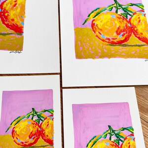 May include: Four prints of a painting depicting two oranges with green leaves on a pink background. The oranges are painted in a stylised way with yellow, orange, and red hues. The background is a light pink colour. The prints are signed "Lilibet".