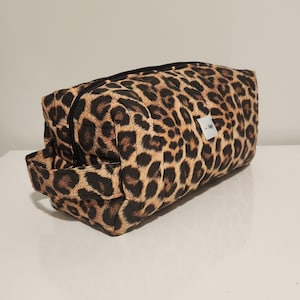 Könnte beinhalten: Eine rechteckige Kosmetiktasche mit Leopardenmuster, einem schwarzen Reißverschluss und einem kleinen Griff an einer Seite. Die Tasche ist beige mit braunen Flecken und einem kleinen weißen Etikett.