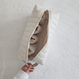 May include: A rectangular, quilted, off-white fabric pouch with a zipper closure. The interior is lined with a beige and white gingham check pattern and features multiple internal fabric dividers for organization, with a small handle on one end.