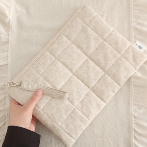 May include: A rectangular, quilted beige pouch featuring a subtle floral pattern, a top zipper closure, and a matching wrist strap. A small white fabric tag on the side displays the text "Saye" with a small "x" symbol printed in black ink.