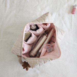 May include: A quilted beige travel bag with a zipper closure, featuring a light pink interior lining with small dark dots. The bag contains a hair styling tool, a round hair brush, and a small hair claw clip. A brown flower charm is attached.
