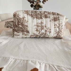 May include: A rectangular toiletry bag with a brown and white tree pattern. The bag has a handle on one side and is placed on a cream-colored linen runner with a ruffled edge. A vase of flowers sits in the background.