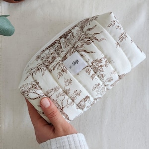 May include: A white and brown quilted cosmetic bag with a toile pattern. The bag has a zip closure and a small label that reads "Saye". The bag is rectangular and has a padded texture.