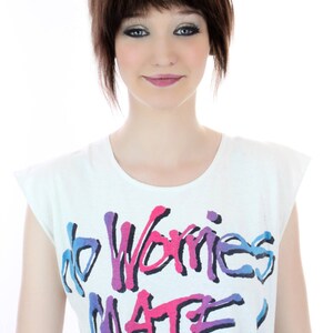 Peut inclure: Un t-shirt blanc avec un graphisme color&eacute; qui dit "no worries mate!" Le graphisme est de style graffiti avec des couleurs rose, bleu et violet.
