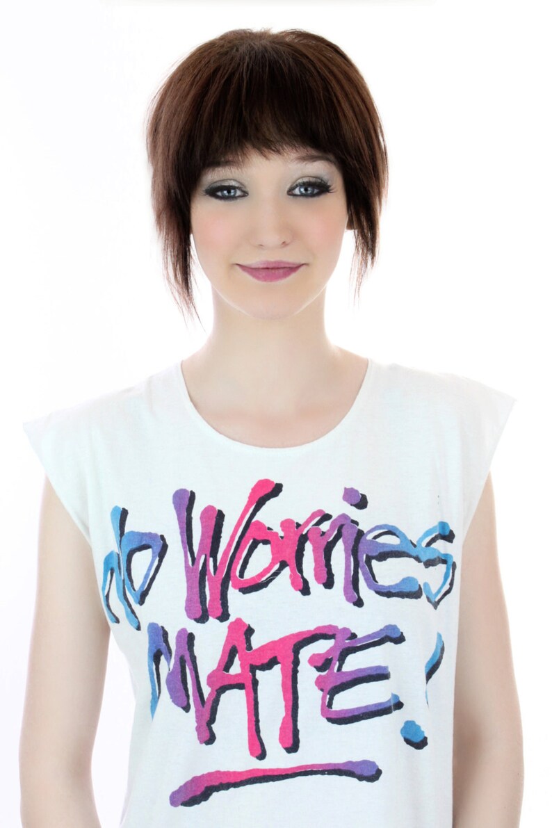Peut inclure: Un t-shirt blanc avec un graphisme color&eacute; qui dit "no worries mate!" Le graphisme est de style graffiti avec des couleurs rose, bleu et violet.