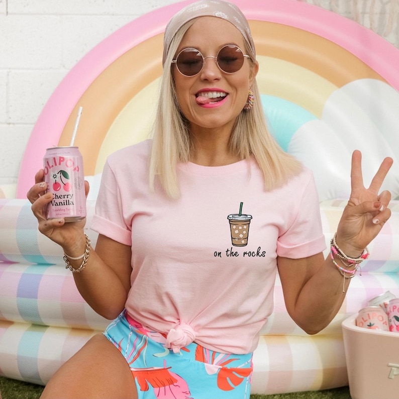 May include: A woman wearing a pink t-shirt with the text "on the rocks" and a drawing of a coffee drink. She is holding a can of cherry vanilla soda and giving a peace sign.