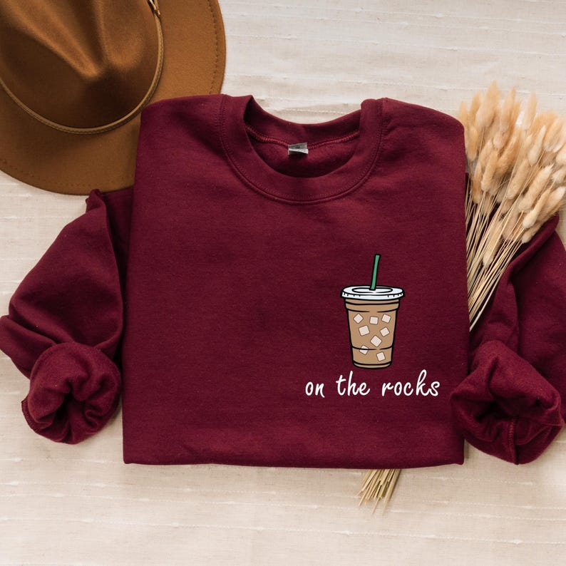 May include: A maroon sweatshirt with a white graphic of a coffee drink with ice cubes and a straw. The text "on the rocks" is printed below the graphic.