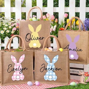 Peut inclure: Sacs fourre-tout en toile de jute avec des motifs de lapin dans des motifs vichy jaunes, roses et bleus. Chaque sac est personnalisé avec un nom en écriture cursive. Les sacs sont remplis d'œufs de Pâques et de verdure.