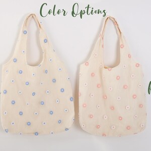 K&ouml;nnte beinhalten: Zwei Tragetaschen mit Blumenstickerei. Die Tasche links hat blaue Blumen und die Tasche rechts hat rosa Blumen. Der Text "Color Options" befindet sich oben im Bild. Der Text "Blue" befindet sich links und der Text "Pink" befindet sich rechts.