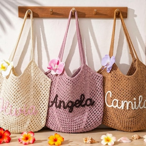 Puede incluir: Tres bolsos de ganchillo colgados en un perchero de madera. El bolso crema lleva el nombre Olivia, el bolso malva lleva el nombre Angela y el bolso marr&oacute;n lleva el nombre Camila. Cada bolso est&aacute; adornado con una flor de orqu&iacute;dea artificial.