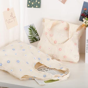 K&ouml;nnte beinhalten: Zwei beigefarbene Einkaufstaschen mit gestickten Blumenmustern. Eine Tasche hat blaue Blumen und die andere hat rosa Blumen. Die Taschen befinden sich auf einem wei&szlig;en Regal mit B&uuml;chern und anderem Dekor.