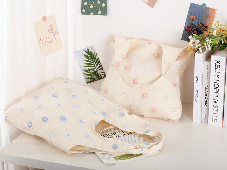 K&ouml;nnte beinhalten: Zwei beigefarbene Einkaufstaschen mit gestickten Blumenmustern. Eine Tasche hat blaue Blumen und die andere hat rosa Blumen. Die Taschen befinden sich auf einem wei&szlig;en Regal mit B&uuml;chern und anderem Dekor.